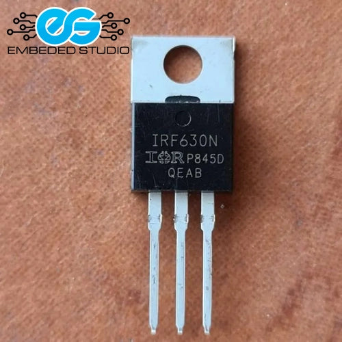 An IRF630N MOSFET transistor in a TO-220 package, featuring a black plastic heat sink with white printed text and a silver metal heat spreader, resting on a brown textured surface.