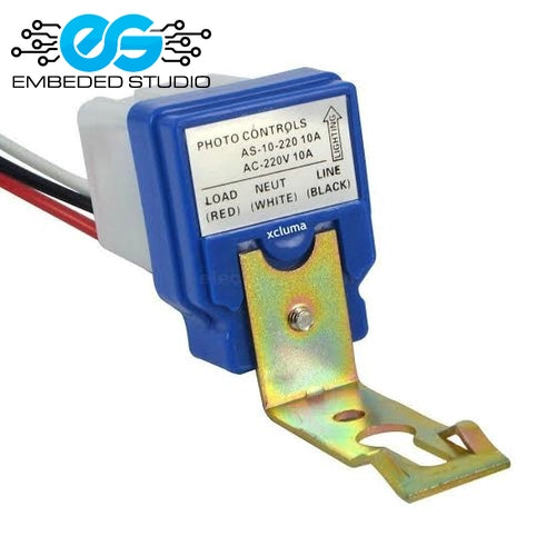 A blue automatic LDR 220V photo control light switch module with a metal mounting bracket, showing wiring labels and the Embeded Studio logo in the top left corner.